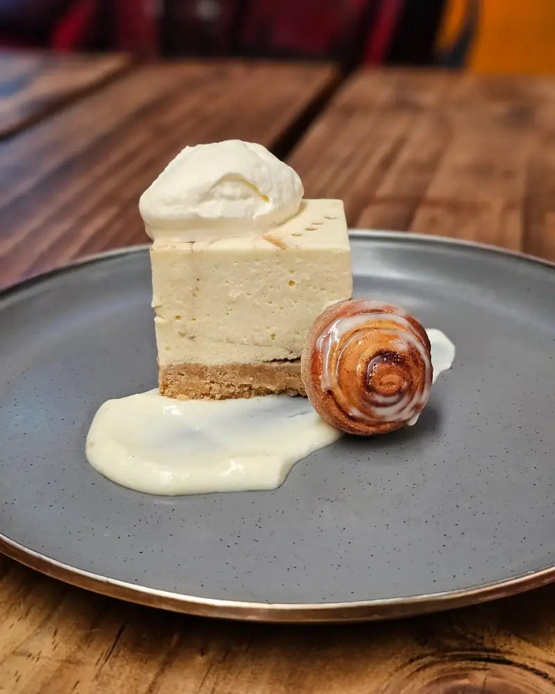 A slice of baked cheesecake with a cinnamon scroll and creme anglaise on a grey plate