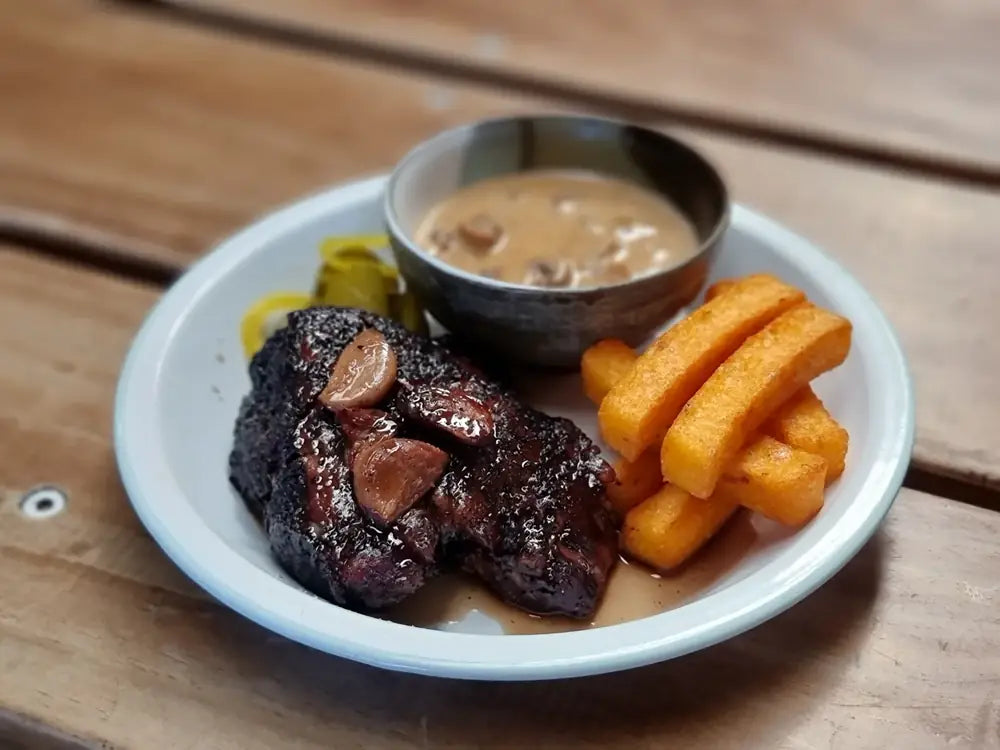 A glazed beef short rib with curly fries, pickles and a pot of gravy on an enamel plate