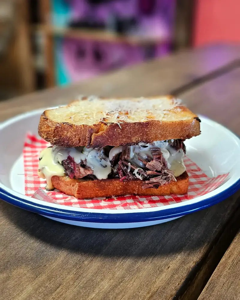A grilled brisket and cheese toastie on an enamel plate lined with gingham paper