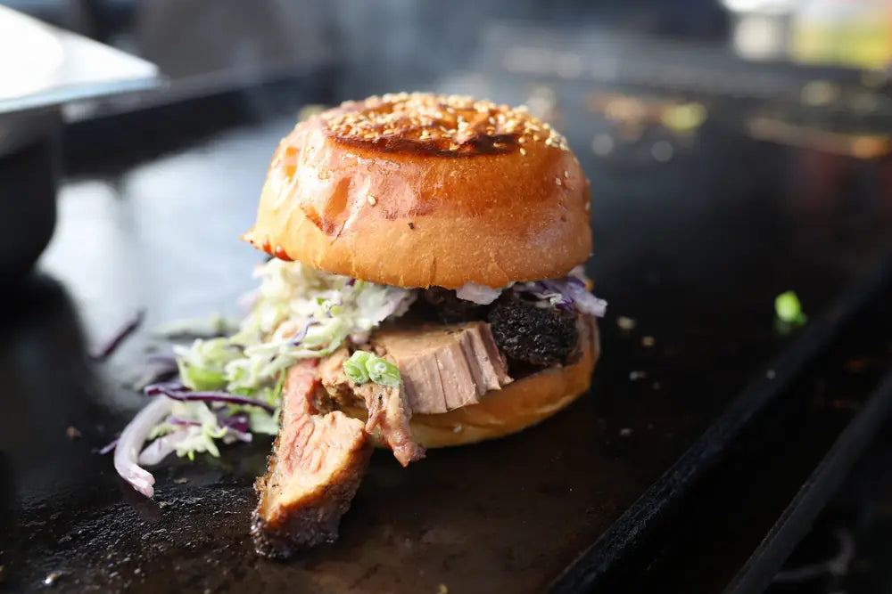 A brisket burger with red cabbage slaw in a brioche bun, resting on the flat top grill