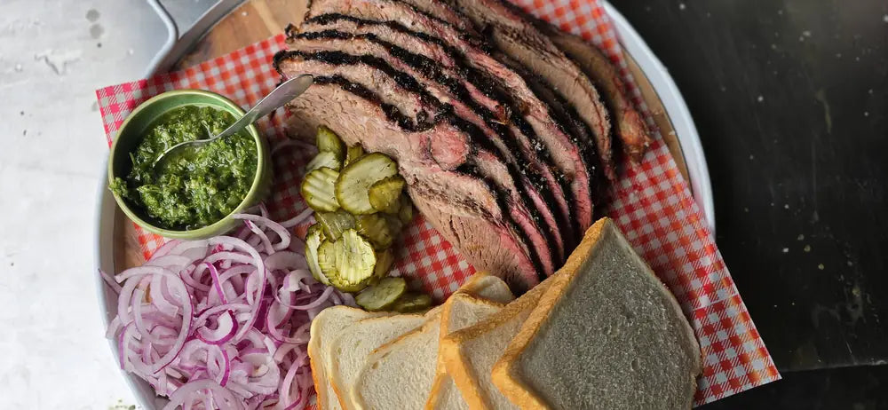 Overhead plate of sliced brisket with chimichurri, dill pickles, pickled red onion and white bread