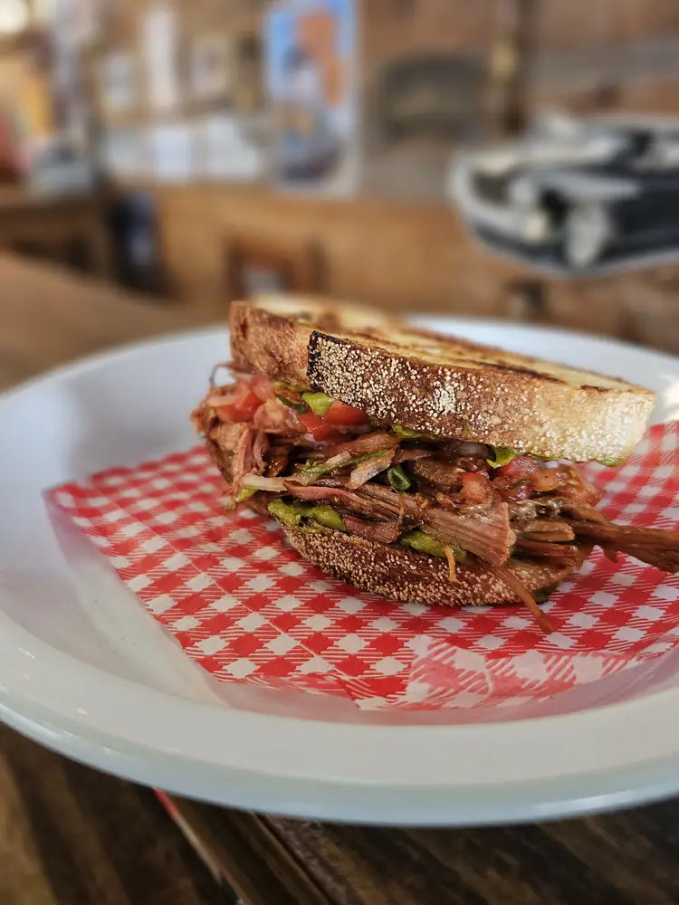 A sliced brisket sandwich with pickles on toasted sourdough, served on gingham paper