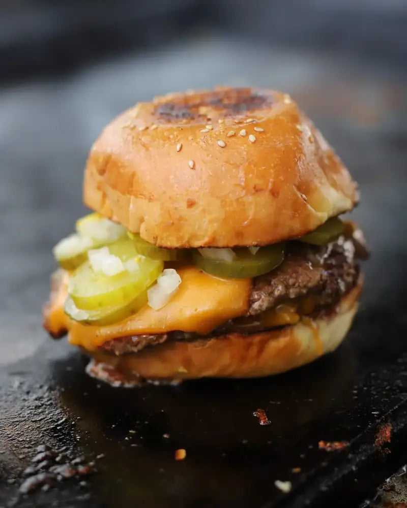 A cheeseburger with pickles and onion in a sesame bun, cooking on the flat top