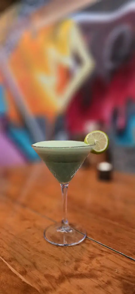 A green cocktail with a lime wheel on the bar, against the mural wall