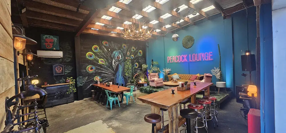 The Peacock Lounge in daylight: the peacock mural, the chandelier, a long communal table and red stools
