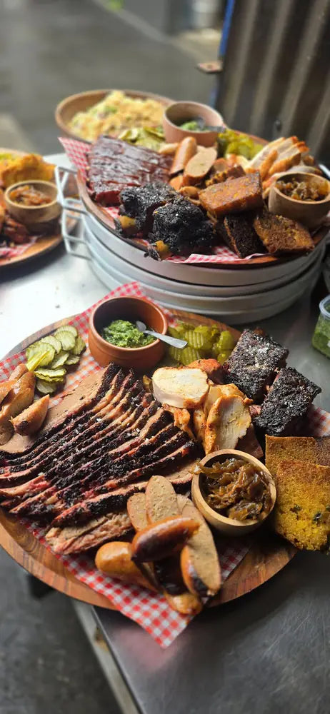 Two loaded sharing platters of brisket, burnt ends, pickles and sides waiting on the pass