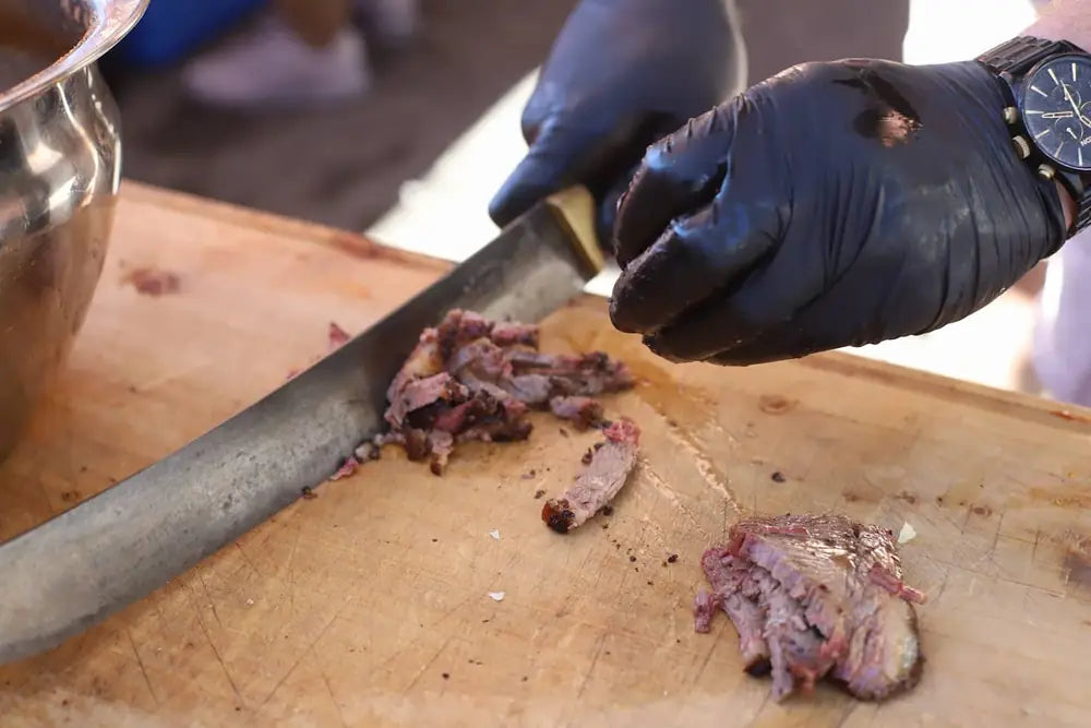 Gloved hands slicing smoked brisket on a wooden board with a carving knife
