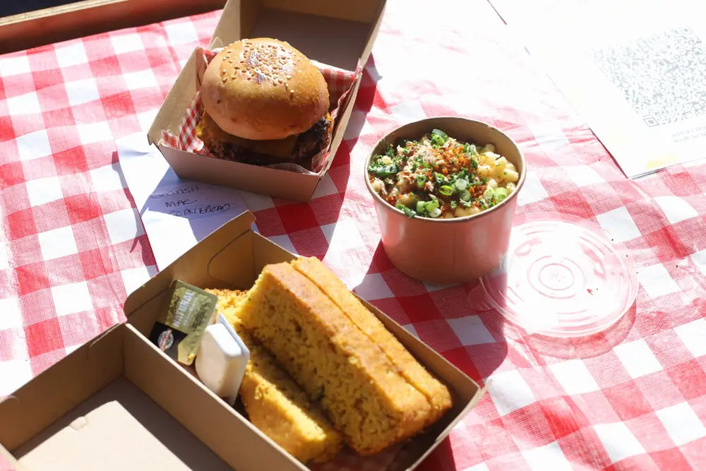 A takeaway box holding a brisket slider, a wedge of cornbread and a cup of corn salad on a red gingham cloth