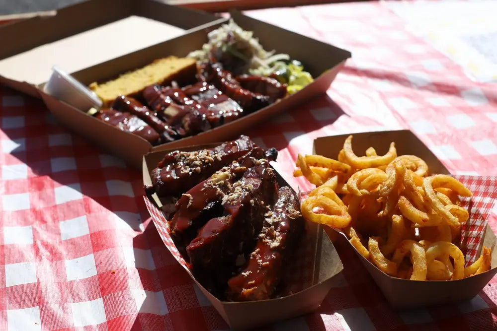 Takeaway trays of glazed pork ribs, cornbread and curly fries on a red gingham cloth