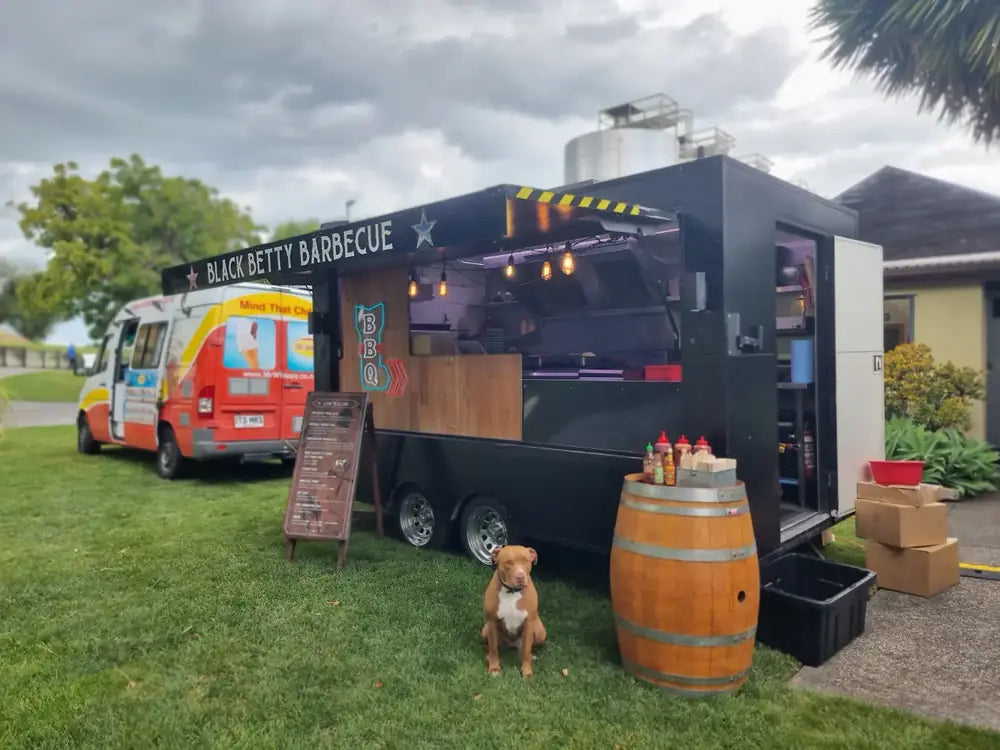 The Black Betty Barbecue food truck and van set up on grass with a whisky barrel and a dog out front