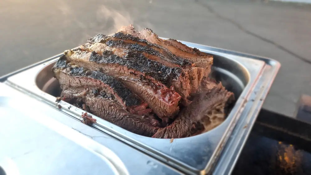 A whole smoked beef brisket with a dark peppery bark, resting in a steel tray